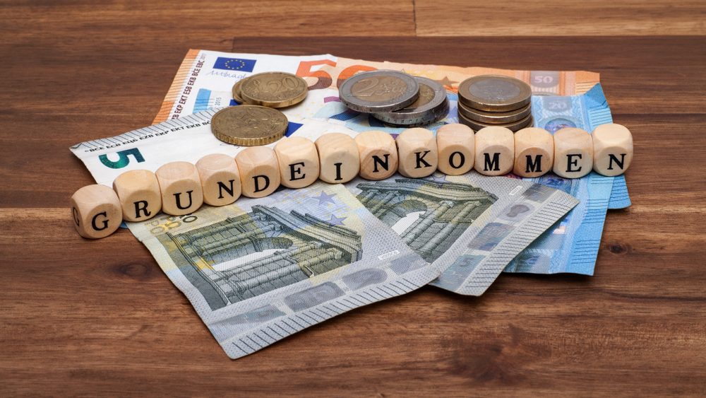 Geldscheine und Münzen, auf denen Holzwürfel mit dem Text "Grundeinkommen" liegen
